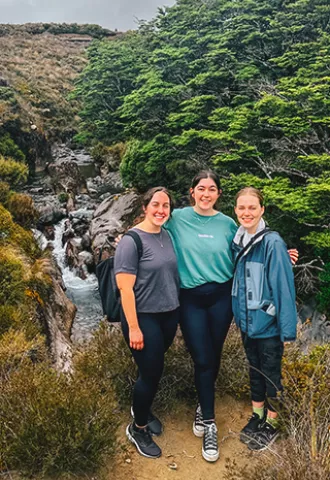 Three people smile together for a photo in front of a small creek.