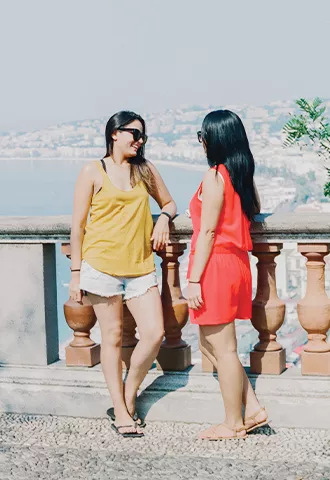 Two people talking on a stone fence with a view of a coastal city behind them.