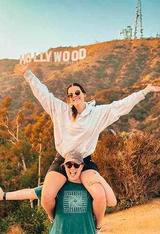 Person sitting on another’s shoulders with their hands in the air in front of the Hollywood sign in Los Angeles.
