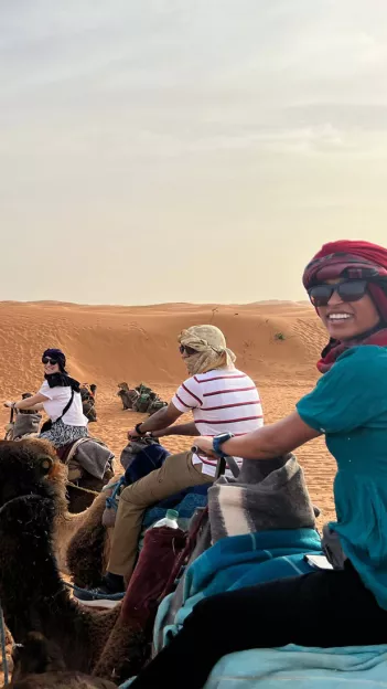 Three woman smiling, wearing sunglasses and cheches while riding camels in the desert.