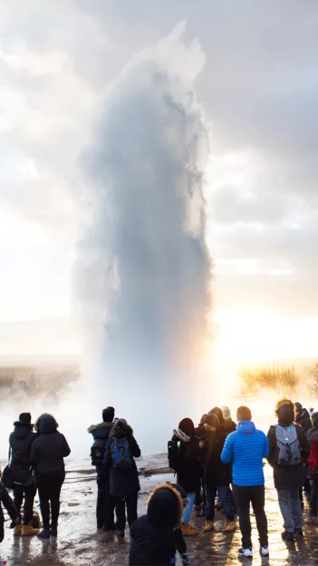 People gather around erupting geyser in Iceland while wearing winter clothing.