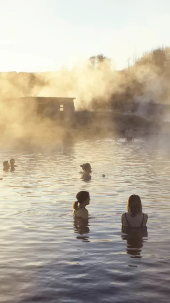 People soak in the waters of the Secret Lagoon in the village of Fludir with steam in the air the sun streaming in.