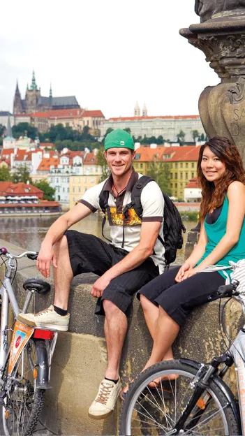 Man and woman on a bike tour, sitting on the Charles Bridge with the Prague Castle in the background.
