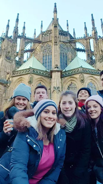 Seven people smile in front of St. Vitus Cathedral in Prague. They are wearing winter jackets and beanie hats.