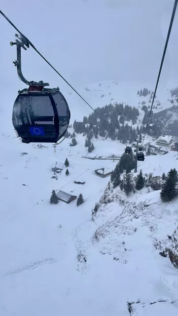  Gondola coming up a snow covered mountain with green trees and snow capped buildings below.