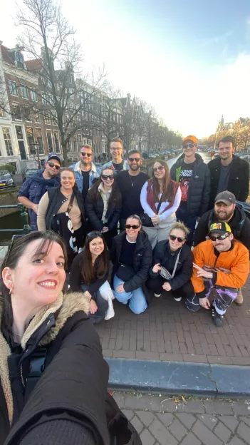 Person taking selfie with group posing together on a canal bridge in Amsterdam.