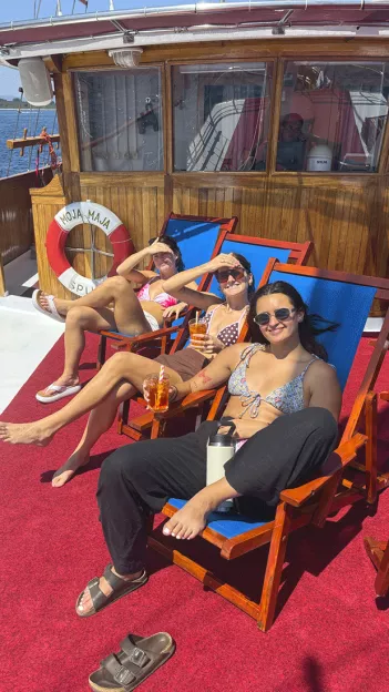 Three people sit smiling in deck chairs at the front of a sailing boat. They wear swim suits and have sunglasses on.