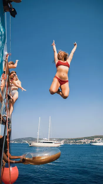 Person poses with their hands up mid air after jumping from sail boat into ocean. Second person holds out floating ring for them to grab.