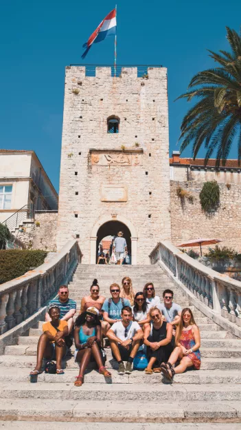 Group of people sitting on stone steps in front of the Revelin Tower. The Croatian flag flies from a flagpole on top of the tower.