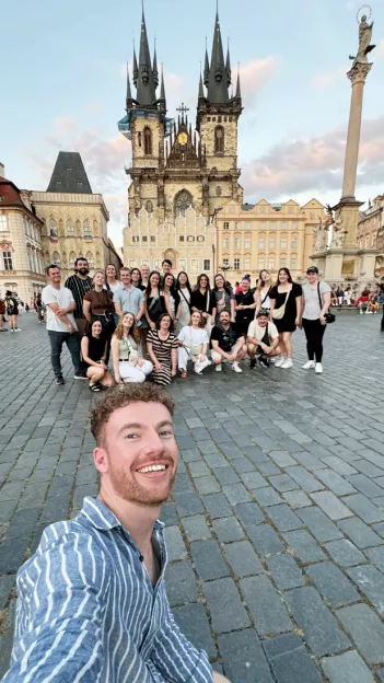 Man takes selfie with group of people behind him, in front of the Church of Our Lady before Týn in Prague.