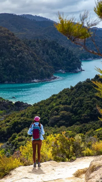 Woman wearing activewear stands on ledge of hill looking out over body of water in New Zealand.