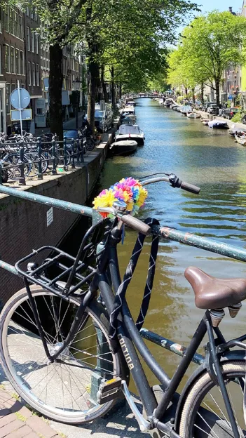 Black bike with colourful faux flowers attached to the frame sits locked to the barrier of a canal bridge in Amsterdam.