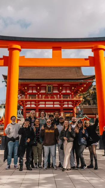 Group smiling and making peace signs with their hands in front of Fushimi Inari Taisha in Kyoto, Japan.