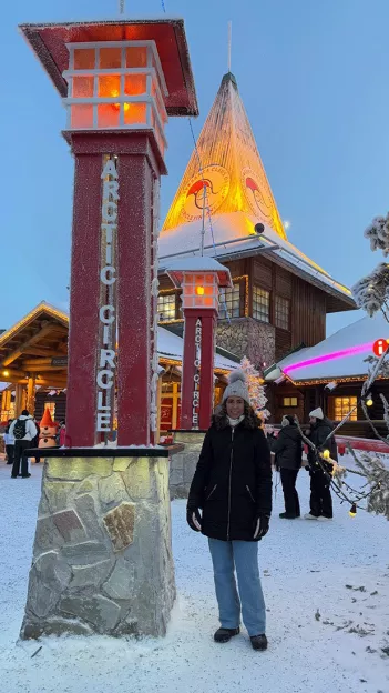 Person wearing winter clothing stands next to a sign for the Arctic Circle.