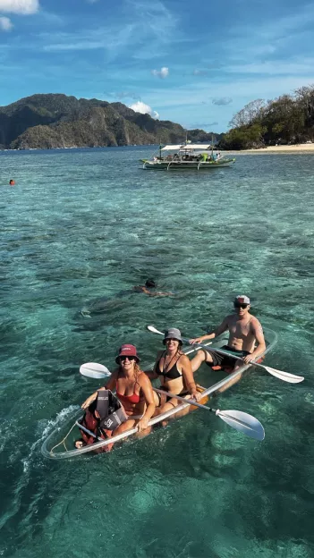 Three people sit in a clear kayak in the blue waters of the Philippines.