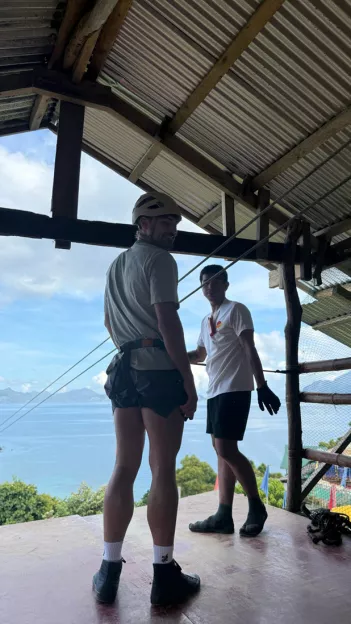 Man smiles while wearing helmet and harness with zipline wire running to his right in a decline.