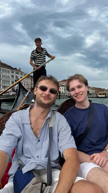 Two people smiling together on a gondola boat in Venice with a gondolier behind them with an ore.