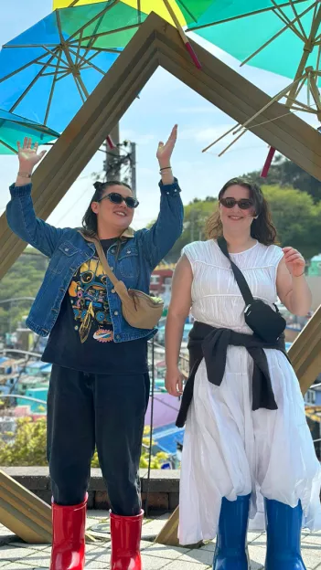 Two women stand in front of an art installation of a slanted picture frame and coloured umbrellas.