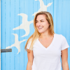 Smiling blonde woman wearing a white top. She stands in front of a blue wall with white birds on it