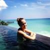 Smiling, blonde woman standing at the edge of an infinity pool with the ocean in the background