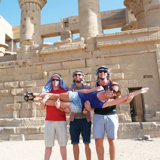 Three men wearing keffiyes and sunglasses, hold up woman posing for picture in front of stone structure.