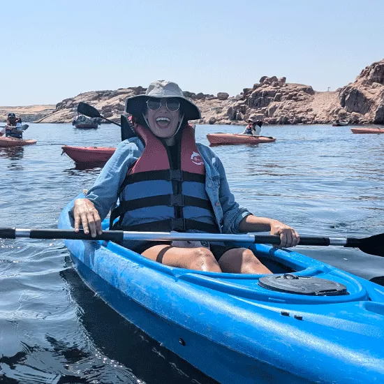  Person sits inside kayak smiling while wearing a sun hat, sun glasses and a life jacket.