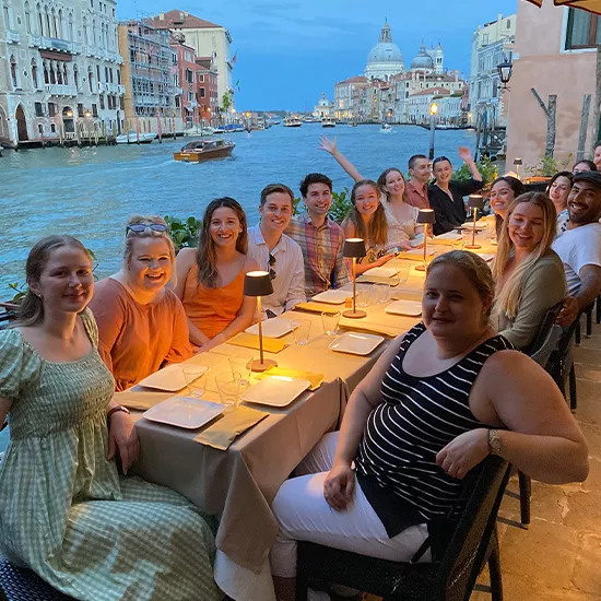 A Topdeck tour group smiles for the camera while eating dinner, with a view of the iconic Venice Canal behind them.