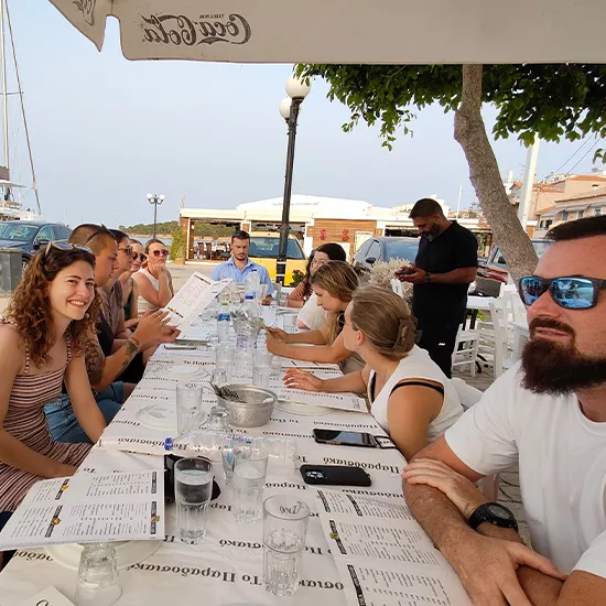 Group of people dining outdoors at a Greek-style taverna located near a marina.