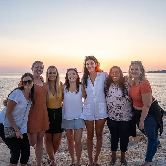 Seven people stand together smiling on a beach in the Greek islands.