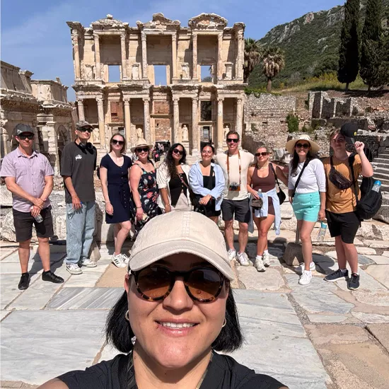 Person smiles while taking a selfie with a group of people posing behind them. In the background is the Library of Celsus in the ancient city of Ephesus.