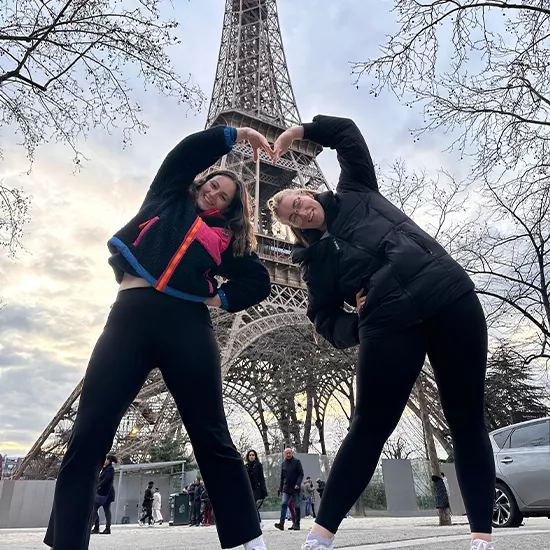 Two Topdeck travellers posing for a photo in front of the Eiffel tower in Winter, using their arms and torsos to make a heart symbol.