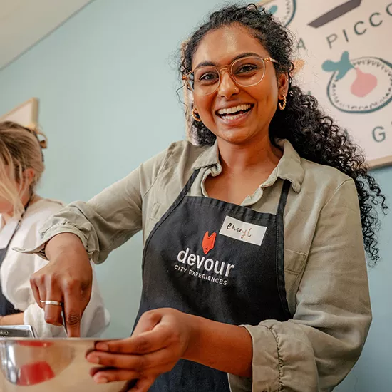 Person smiling while stiring something within a metal bowl. They wear an apron with a name tag.