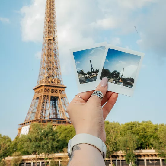 Hand holds two polaroid style pictures up to the sky with the Eiffel Tower in the background.