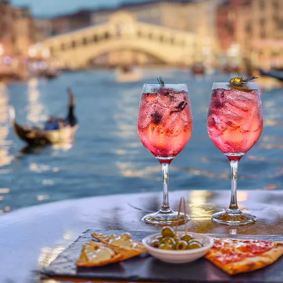 Two glasses of Aperol spritz, slice of pizza and small bowl of olives on a table next to the Grand Canal in Venice.