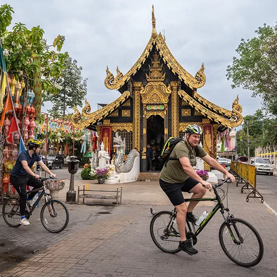 Two people on bikes passing by a traditional Thai temple.